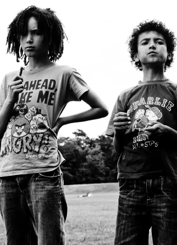 A black and white photograph of two young people (s2, s3) standing outdoors. On the left is a person with shoulder-length dreadlocks wearing an Angry Birds t-shirt that reads AHEAD, MAKE ME ANGRY with cartoon bird characters. On the right is a person with curly hair wearing a Charlie Brown All-Stars t-shirt featuring Peanuts characters. Both are wearing jeans and appear to be standing on a path or open area with trees visible in the background. The image has an artistic, portrait-style composition shot from a low angle looking up at the subjects.