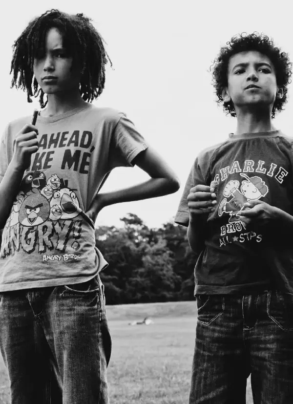 A black and white photograph of two young people (s2, s3) standing outdoors. On the left is a person with shoulder-length dreadlocks wearing an Angry Birds t-shirt that reads AHEAD, MAKE ME ANGRY with cartoon bird characters. On the right is a person with curly hair wearing a Charlie Brown All-Stars t-shirt featuring Peanuts characters. Both are wearing jeans and appear to be standing on a path or open area with trees visible in the background. The image has an artistic, portrait-style composition shot from a low angle looking up at the subjects.