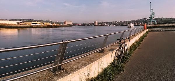 A touring bicycle parked against metal railings beside the River Medway. The river stretches into the distance with industrial buildings and structures visible on the opposite bank. The path appears to be part of a waterside cycling route with modern safety barriers.