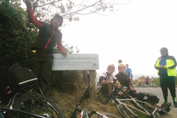 A group of cyclists taking a rest break at Ditchling Beacon with their bicycles, where several people are sitting on the ground while one person stands with arms raised in celebration next to a welcome sign, with misty or foggy conditions visible in the background