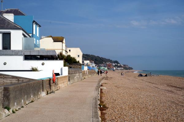 A seaside promenade lined with modern and older buildings, people walking along the pathway, a shingle beach stretching out to the right, and the sea under a clear blue sky.