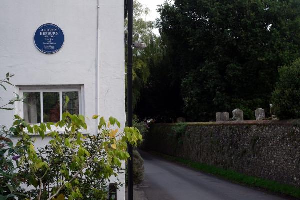 A white house with a blue plaque dedicated to Audrey Hepburn, viewed from an angle showing a leafy plant in front and a narrow village lane running beside an old stone wall with grave markers visible over the top.