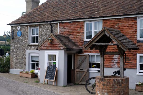 A rustic country pub with a red and flint facade, a blue plaque above the door commemorating Ian Fleming, a wooden well in the foreground, a bicycle leaning nearby, and a chalkboard displaying opening hours.