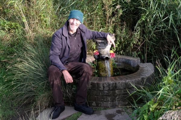 A man sits smiling beside the historic Harty Ferry artesian well at Oare Marshes nature reserve in Kent. He wears a dark green hat, dark jacket, and dark trousers while seated on the stone structure surrounding the well. The well features a traditional design with weathered stone construction and a metal spigot from which water flows into a collection basin. Pink flowers have been placed decoratively on the wellhead, adding a cheerful touch to this important local landmark. Tall marsh grasses and vegetation surround the well, creating a natural setting typical of the Oare Marshes wildlife reserve. This artesian well, originally drilled in 1900 to a depth of 76 meters, provides mineral-filtered water from the same chalk aquifer that supplies Shepherd Neame brewery in Faversham. The well ceased flowing in late 2018 but was successfully restored in 2024 through funding from Swale Borough Council and repair work by Kent Wildlife Trust.