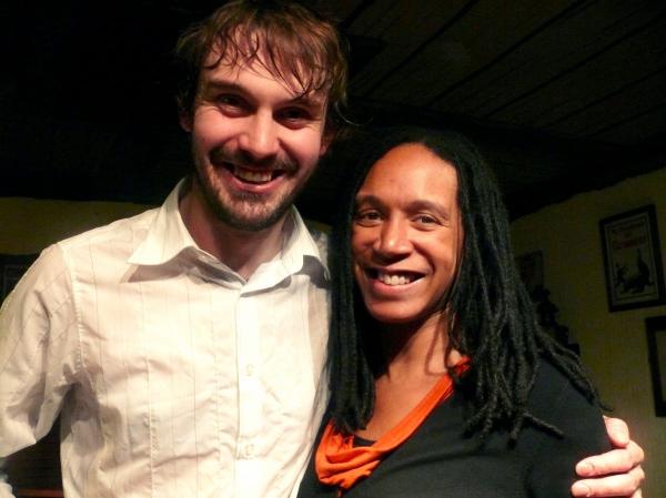A smiling man with damp hair wearing a white collared shirt poses with his arm around a woman with long dark hair wearing a black top with an orange accent, both appearing happy and relaxed in what looks like an indoor setting with framed artwork visible on the walls behind them