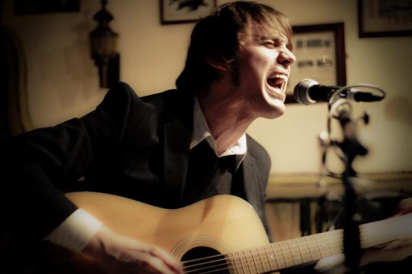 A young blonde man with shoulder-length hair singing passionately into a microphone while playing an acoustic guitar in what appears to be a recording studio or intimate performance space with framed pictures on the walls behind him