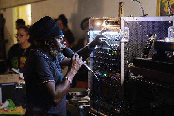 A person operating a large sound system control panel at an indoor event, holding a microphone with one hand and adjusting audio equipment with the other, surrounded by racks of illuminated audio gea