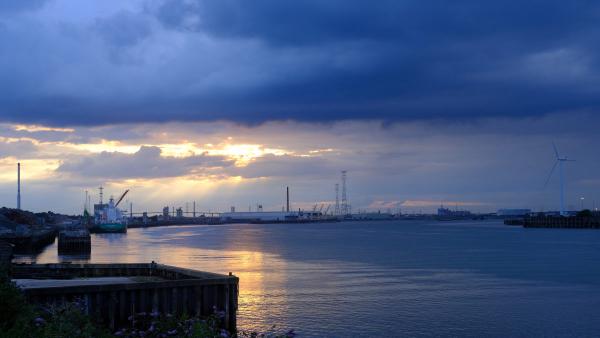 A river or estuary scene at sunset with dramatic clouds, the water reflecting the fading sunlight, and silhouettes of industrial infrastructure such as cranes, warehouses, and a wind turbine along the shoreline.