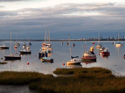 A harbor scene at dusk with numerous small boats and yachts moored in calm water, featuring a cloudy sky with warm evening light and grassy salt marshes in the foreground.