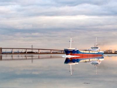 A serene waterscape showing a blue and red boat moored near a long wooden pier or bridge, with perfect mirror reflections of both the vessel and cloudy sky in the still water.