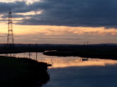 A dramatic sunset landscape showing a winding waterway reflecting cloudy skies, with an electrical transmission tower silhouetted against the golden and blue evening sky.