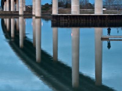 A modern concrete bridge with multiple cylindrical support pillars crossing over calm water, creating perfect reflections of the structure and overcast sky in the still water below. The bridge appears to curve slightly and has a person visible on a small platform or dock near the water's edge.