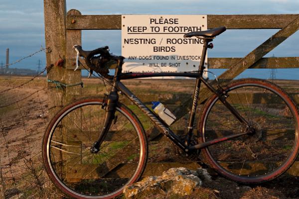 A black touring bicycle leaning against a wooden fence gate with a warning sign about keeping to footpaths and nesting birds, set in a rural countryside location with rolling hills in the background.
