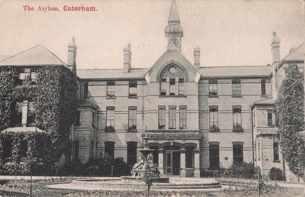 Historic black-and-white photograph of The Asylum, Caterham, showing a large Victorian institutional building with a central clock tower and multiple chimneys. The frontage is symmetrical, with tall windows, ivy climbing the walls, and a central entrance. In front, there is a circular fountain featuring sculpted figures, surrounded by a neatly maintained garden area. The overall appearance is imposing and characteristic of 19th-century institutional architecture.