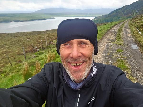 A person stands in the foreground on a grassy verge next to a rough country road, wearing outdoor gear with rolling hills, a large body of water, and cloudy skies visible in the background.