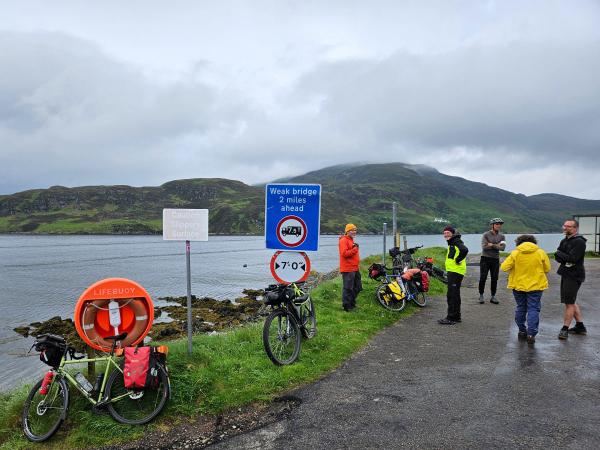 Several bicycles with touring panniers are parked along a roadside next to a waterfront. A group of people, some in bright outdoor jackets, are gathered near road and caution signs. Hills covered in greenery rise across the water under a cloudy sky.