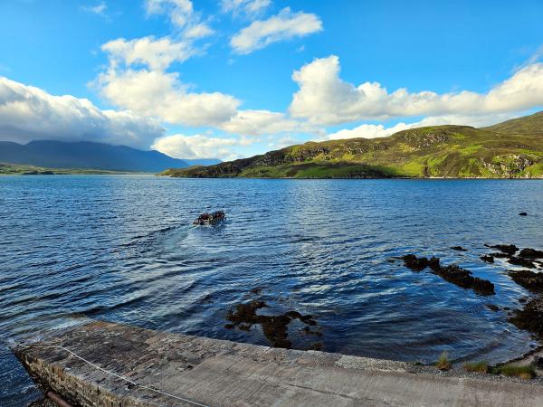 A small metal boat carrying several passengers speeds away from a stone jetty across a wide lake, with green hills and mountains in the background under a sky with scattered clouds.