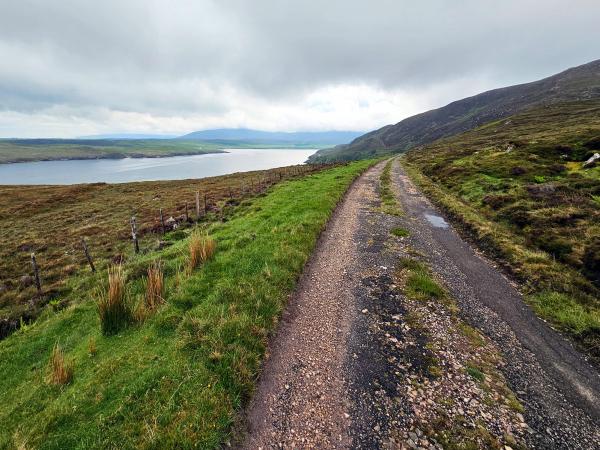 A wide view of rolling green hills and cliffs meets a distant coast and bay. The landscape is rugged and open, the water stretches out to the horizon, and the sky is covered with thick clouds.