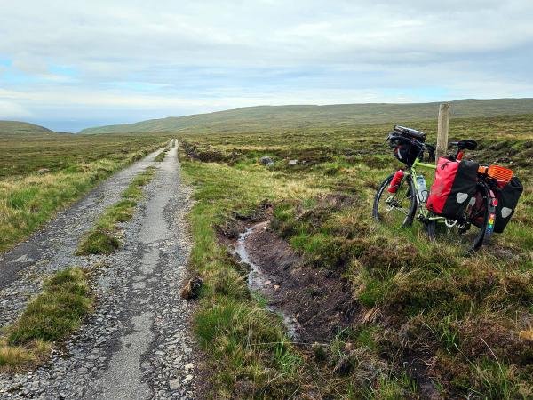 A loaded touring bicycle rests beside a cracked and weathered moorland road that disappears into the horizon. Rolling green hills and distant sea are visible beneath a partly cloudy sky.