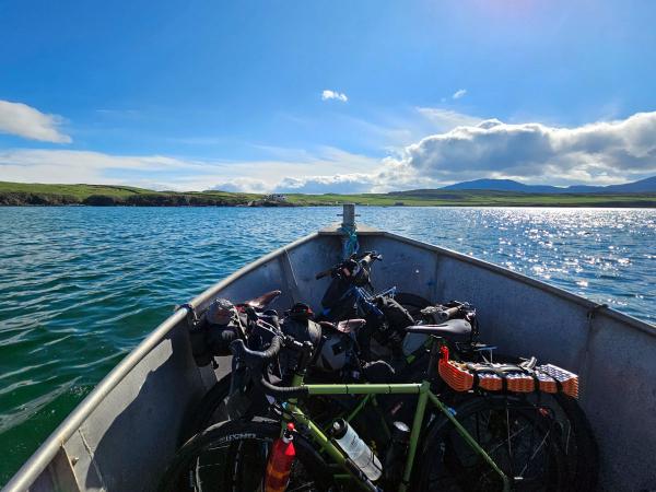 Several bicycles rest in the bow of a small metal boat as it floats across a lake toward green hills under a sunny sky with puffy clouds.