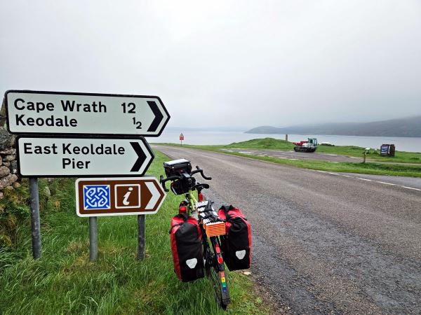 A fully loaded touring bicycle stands next to road signs indicating directions: Cape Wrath 12, Keoldale ½, and East Keoldale Pier. Beyond the signs, a quiet road runs beside a bay with distant hills fading into the mist.