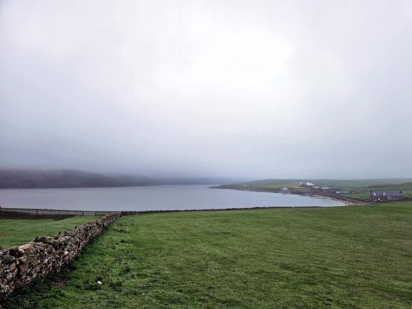 A wide, misty view of a calm bay with green fields in the foreground. A low stone wall runs towards the water, and scattered white houses are visible along the distant shoreline. Hills shrouded in fog rise across the water.