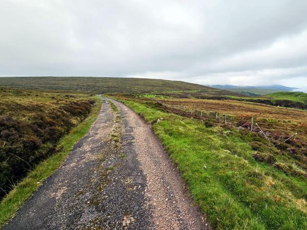 A rough, rural track runs alongside a wire fence overlooking a large body of water and rolling hills. The landscape is lush and green, and the sky is cloudy and grey.
