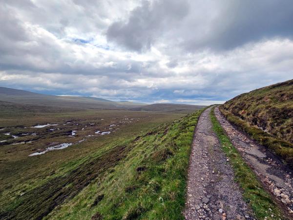 An old gravel road curves along the side of a grassy hill, overlooking expansive, waterlogged moorland below with mountains on the horizon and a dramatic, cloudy sky.