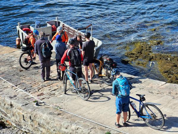 A group of people with bicycles gathers on a sloped stone jetty next to a small metal boat tied up at the water’s edge. Some people wear helmets and backpacks, and the scene is bathed in bright daylight.