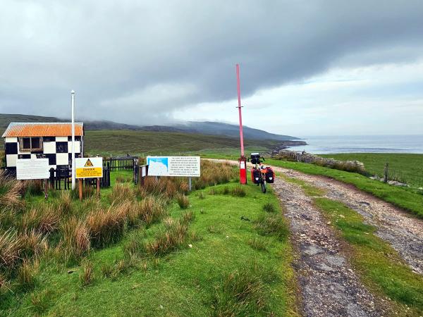 A remote, gravel road leads to a small hut painted in a checkerboard pattern with several warning and informational signs. A bicycle loaded with bags is parked by a raised gate beside the road, with grassy fields and the coastline in the distance.