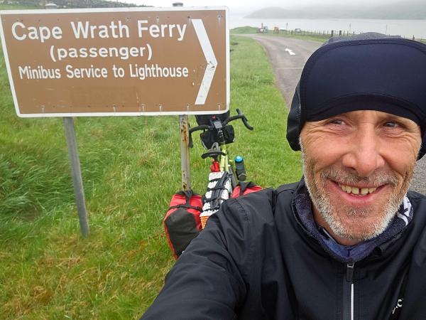 A person stands beside a brown sign that reads Cape Wrath Ferry (passenger) Minibus Service to Lighthouse. Behind them, a loaded touring bicycle leans against the signpost. The grassy verge and a winding road lead down to misty waters in the background.