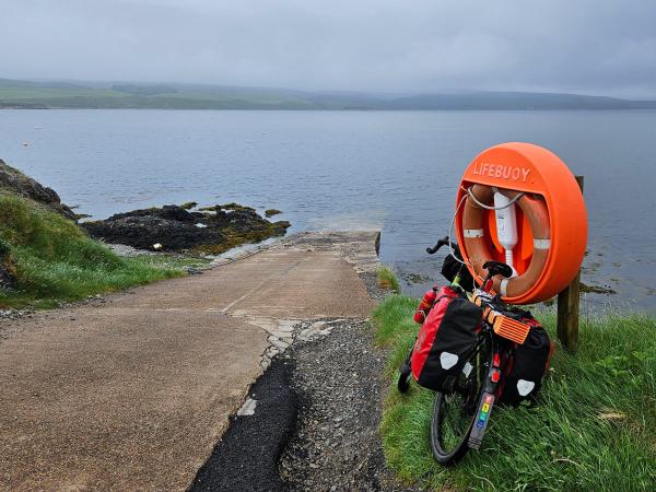 A loaded touring bicycle leans against a wooden post with an orange lifebuoy mounted on it beside a slipway leading down to a calm body of water. The setting is overcast with distant green hills visible across the water.
