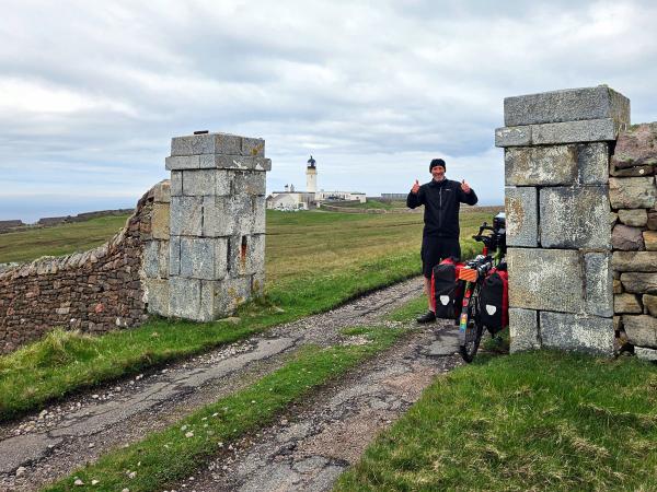 A cyclist stands between two stone gateposts on a rough path giving a double thumbs up, with a lighthouse and low white buildings visible across an open grassy landscape under a cloudy sky. A bicycle loaded with bags is parked nearby.