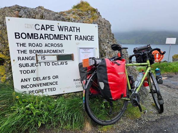 A touring bicycle leans against a large white sign that states Cape Wrath Bombardment Range and provides a warning that the road across the range may be subject to delays. Bright green grass, a rocky outcrop, and water are visible in the background on a cloudy, misty day.