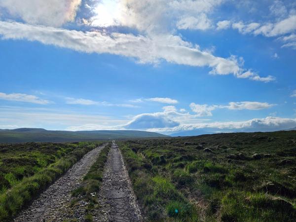 Long straight stony track runs through open moorland covered in rough grass and heather. The sky is bright blue with scattered clouds and the sun shining through, creating a scenic and remote atmosphere.