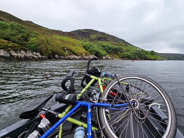 Two loaded touring bicycles lie in a small boat or ferry crossing a scenic body of water. The background features steep, verdant hillside slopes dotted with shrubs and trees beneath a grey sky.