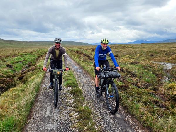 Two cyclists ride sturdy off-road bikes along a narrow, rough path bordered by grass and moorland, under an overcast sky.