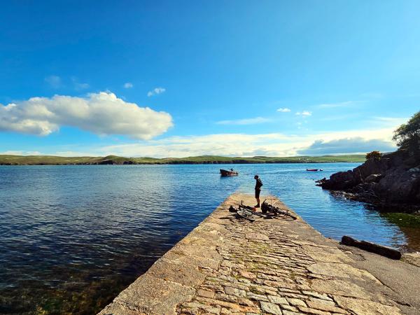 A person stands on a stone pier next to two bicycles, gazing across a calm lake under a bright blue sky with scattered clouds. The surrounding landscape features low, green hills and another small boat is visible on the water in the distance.