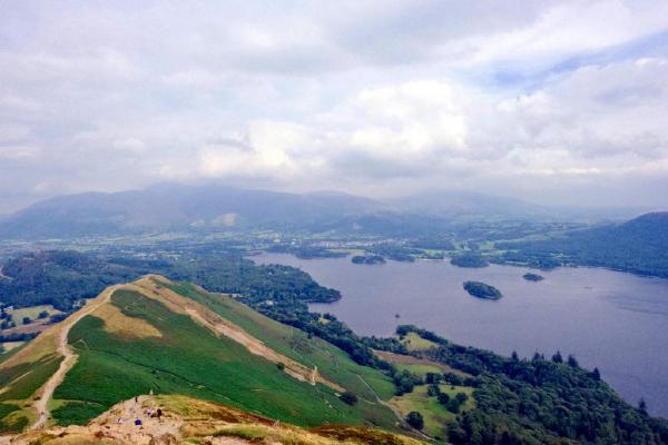 A panoramic view from the top of Cat Bells looking down over Derwent Water, with its wooded islands visible on the calm, grey-toned lake. Rolling fells and patches of forest stretch beyond the water, with distant mountains fading into mist. In the foreground, grass and golden-brown heath cover the slope, and a path winds up toward the summit.