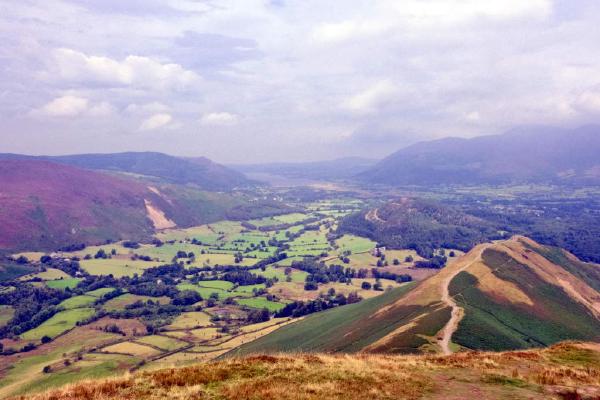 A wide panoramic view from a fell top looking down into a broad valley of patchwork green fields divided by hedgerows and tree lines. Golden-brown grass covers the foreground slope, with a faint path curving down to the right. A steep hill with reddish-purple tones rises on the left, and distant mountains fade into blue-grey haze under a partly cloudy sky.