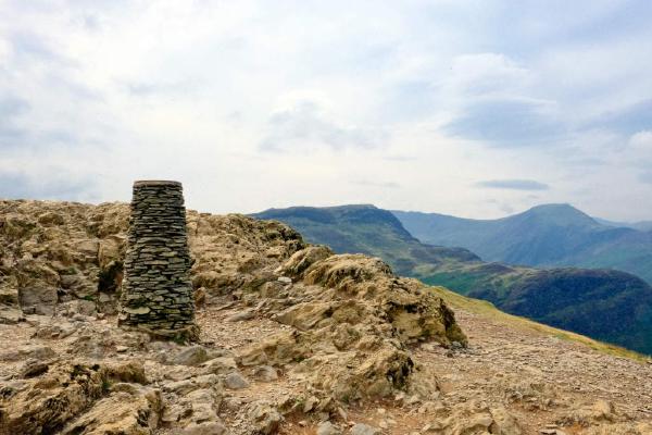 A stone trig point standing on a rocky outcrop of scattered boulders and scree, with a faint path winding down the slope below. Layered fells recede into blue-grey distance under a softly overcast sky, with patches of green vegetation visible in the valley.