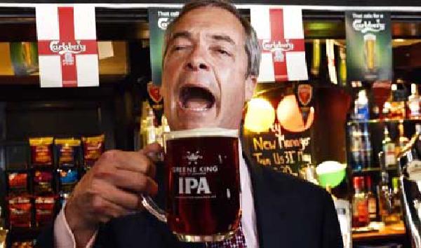 A middle-aged man in a dark suit enthusiastically drinking from a pint glass of beer, appearing to be celebrating. Behind him is a bar setting with various bottles and English flags visible, suggesting this is taken in a pub or similar venue