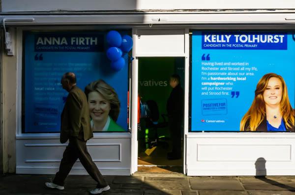 A Conservative Party campaign office storefront with large blue poster displays. The left poster features Anna Firth with text about being a candidate in the Postal Primary. The right poster shows Kelly Tolhurst with quotes about housing and planning policies. A person walks past the storefront in the foreground.