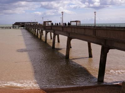 Deal Pier. The last remaining fully intact pleasure pier in Kent. Long concrete pier stretching into the sea with people walking beneath a cloudy sky.