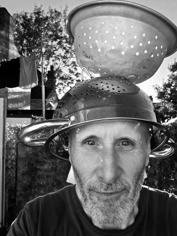 Black and white portrait of a man outdoors wearing two stacked metal colanders as an improvised eclipse-viewing helmet, with dappled shadow patterns projected onto his face by sunlight filtering through the perforations during a partial solar eclipse in Chatham.