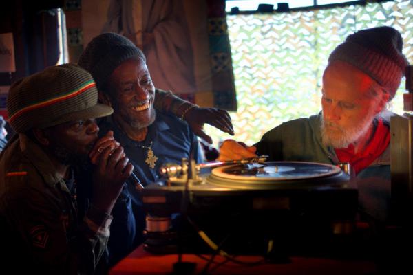 Three people gather closely around DJ equipment and a turntable. One person appears to be selecting or placing a record, while another holds a microphone, and a third stands beside them. All are in a warmly lit room decorated with colorful fabrics.