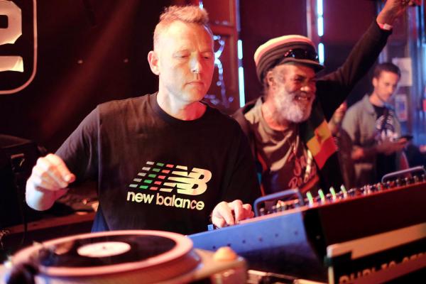 Two people are standing at a DJ booth, one wearing a New Balance T-shirt and the other in a dark jacket and hat with a rasta-colored band, appearing to select or play vinyl records on turntables and mixing equipment in a dimly lit club setting. A third person is visible in the background.