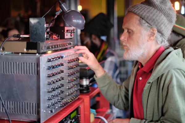 An individual with greying hair and a wool beanie adjusts a large analog audio mixing console, which is equipped with many dials and sliders. The environment suggests a music event with other people nearby, including one with a colorful scarf and hat.