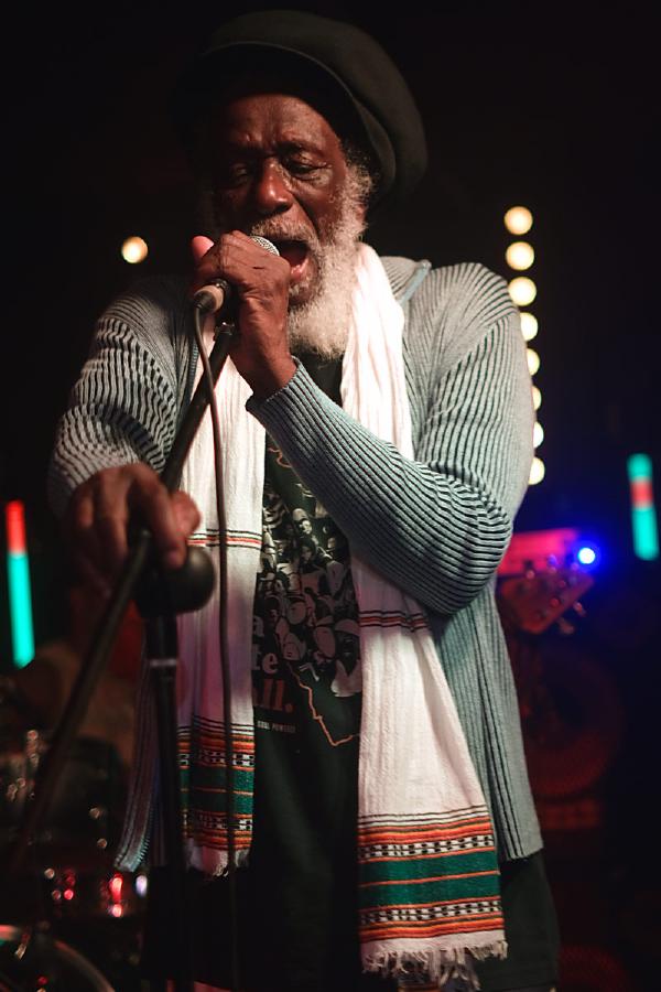 Ashanti Roy (Roydel Johnson) of The Congos performing on stage wearing a striped garment with traditional African-style wrap and dark hat. Colorful stage lights illuminate the background as he sings into a microphone with an expressive gesture.