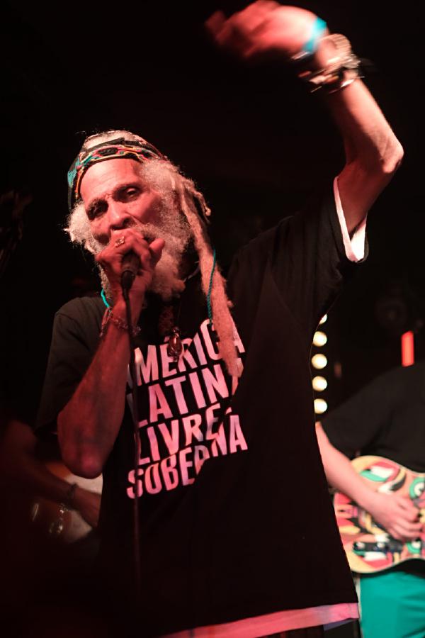 Cedric Myton performing with dramatic lighting, wearing the “AMERICA LATINA LIVRE E SOBERANA” t-shirt and colorful headband. The image captures him mid-performance with his arm raised in a dynamic blur of motion, microphone in hand.